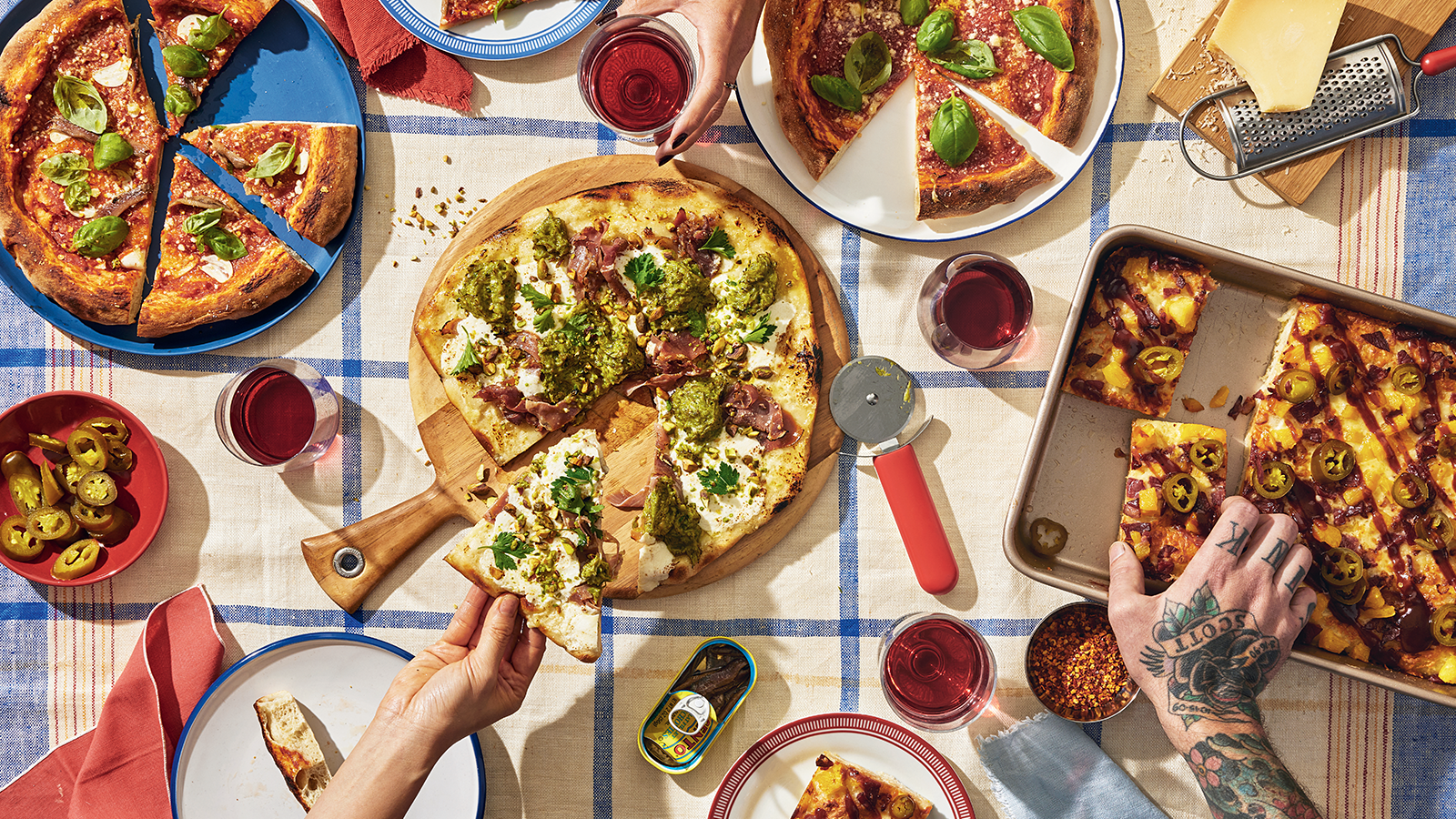 An overhead shot of a table with four different pizzas, wine glasses, tins of anchovies, and hands grabbing slices of pizza.